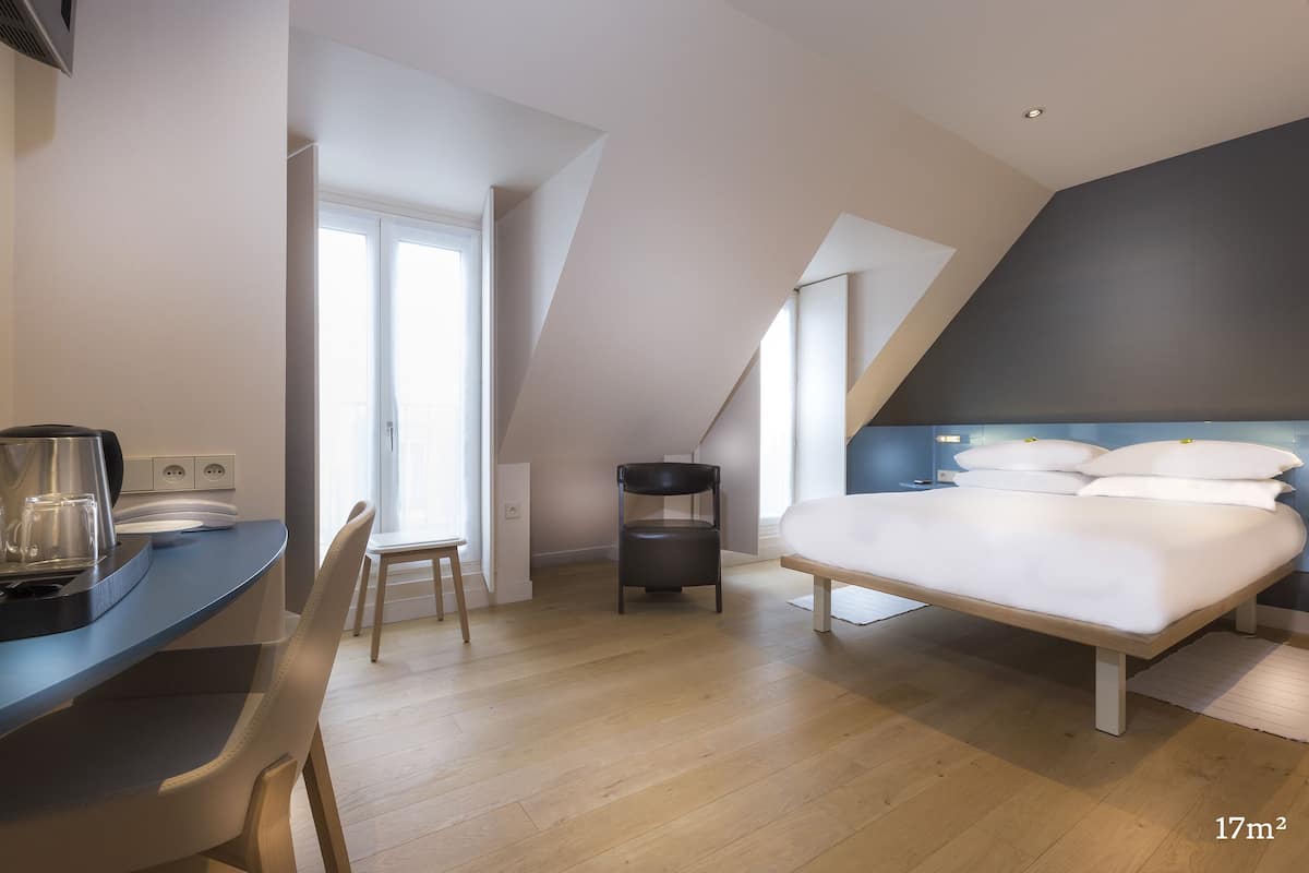 A modern bedroom with a large bed, a desk, and a chair. There is a window providing natural light, and the room has a sloped ceiling.