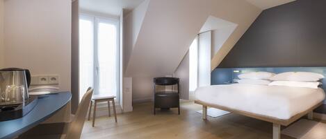 Un dormitorio moderno con una cama grande, un escritorio y una silla. Hay una ventana que proporciona luz natural, y la habitación tiene un techo inclinado.