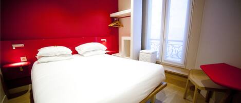 A modern hotel room with a red accent wall, a large bed with white linens, a small bedside table, a red chair, and a window with a balcony view.