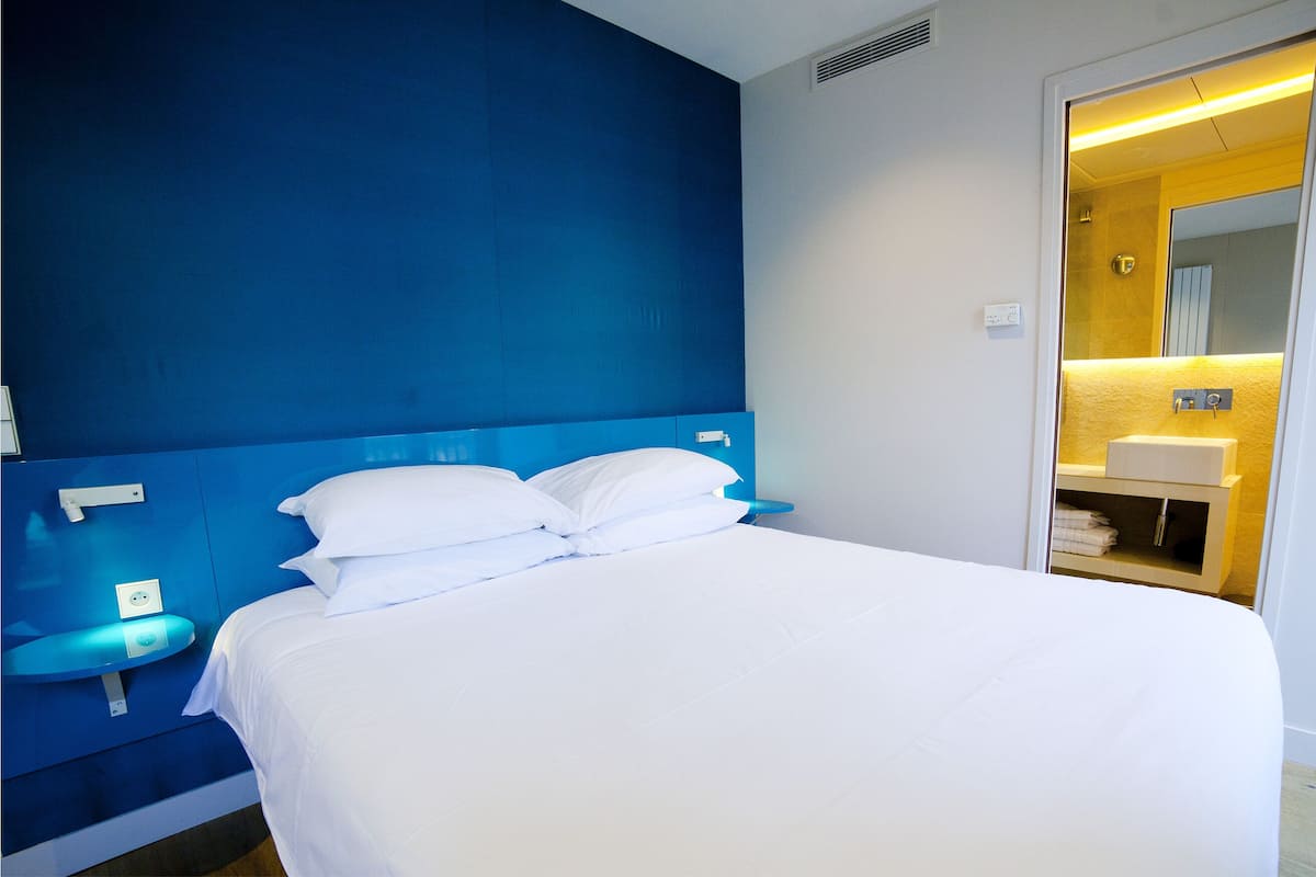 A hotel room with a large bed, a bedside table, a blue headboard, and a bathroom visible through an open door.