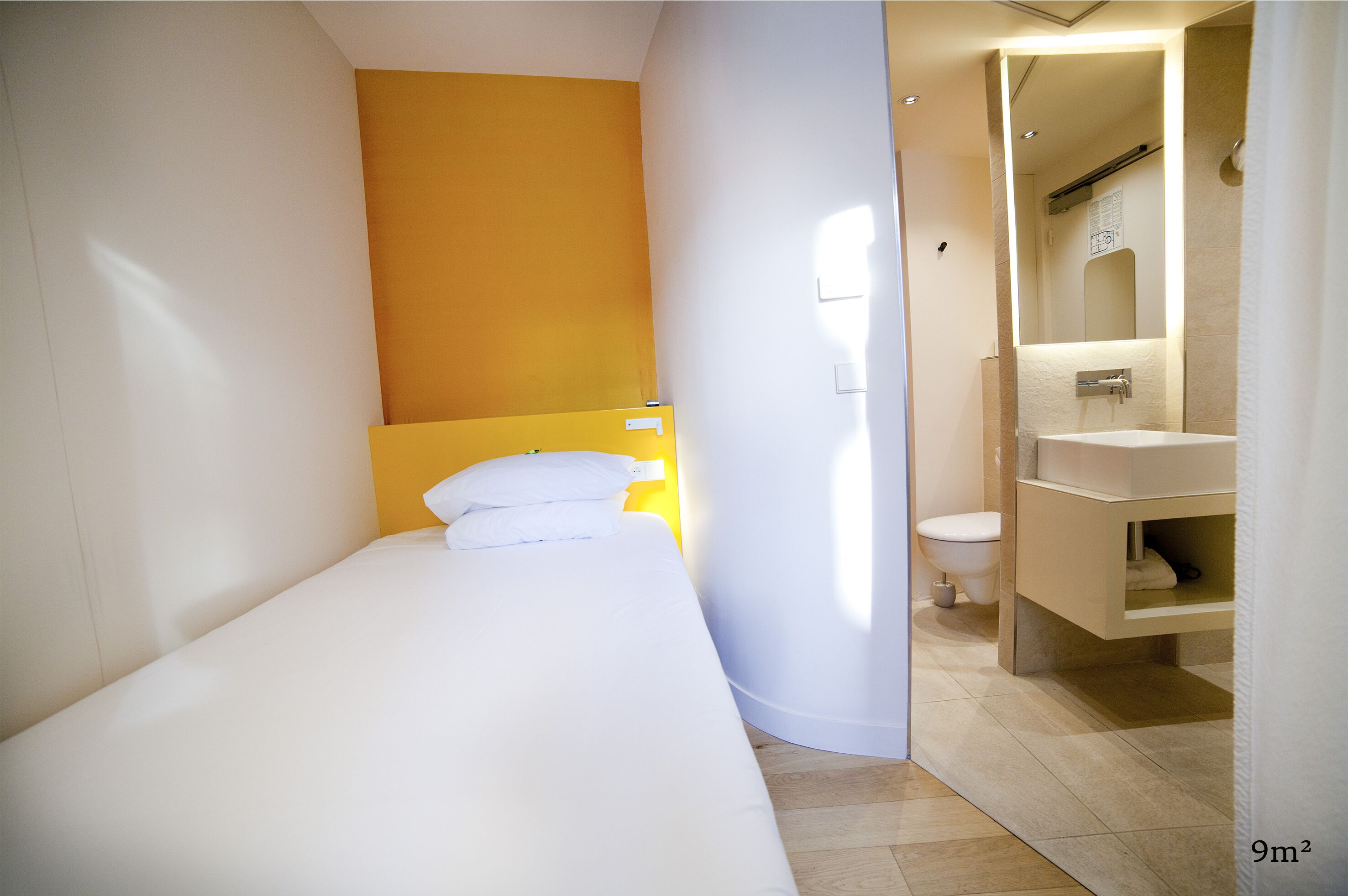 A small, modern room with a bed, a yellow accent wall, a bathroom with a sink and toilet.