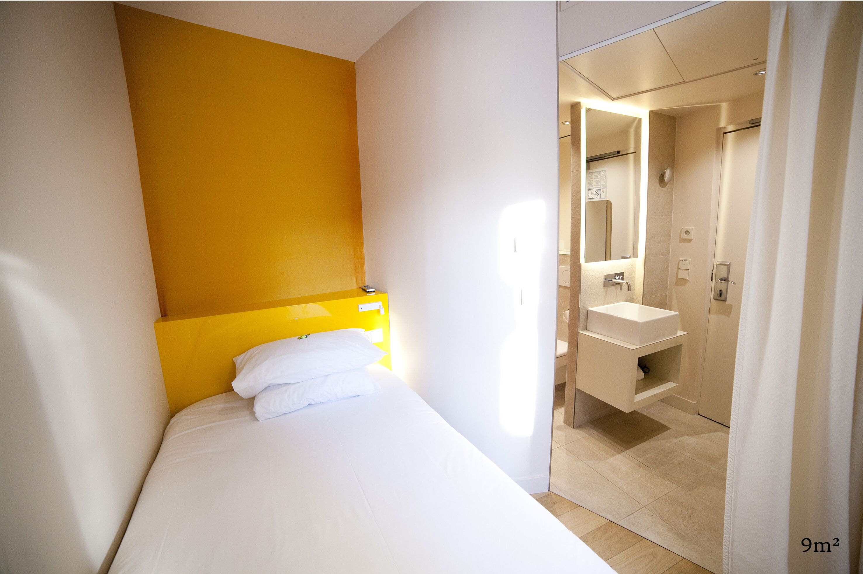 A hotel room with a bed, a yellow headboard, a white wall, a bathroom with a sink and mirror, and a shower.
