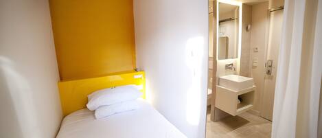 A hotel room with a bed, a yellow headboard, a white wall, a bathroom with a sink and mirror, and a shower.
