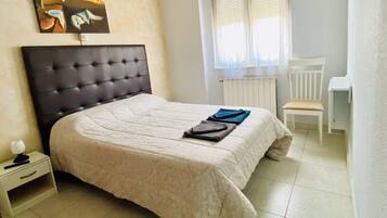 Una cama bien tendida con cabecera, una mesita de noche con lámpara, una silla y una ventana con cortinas.