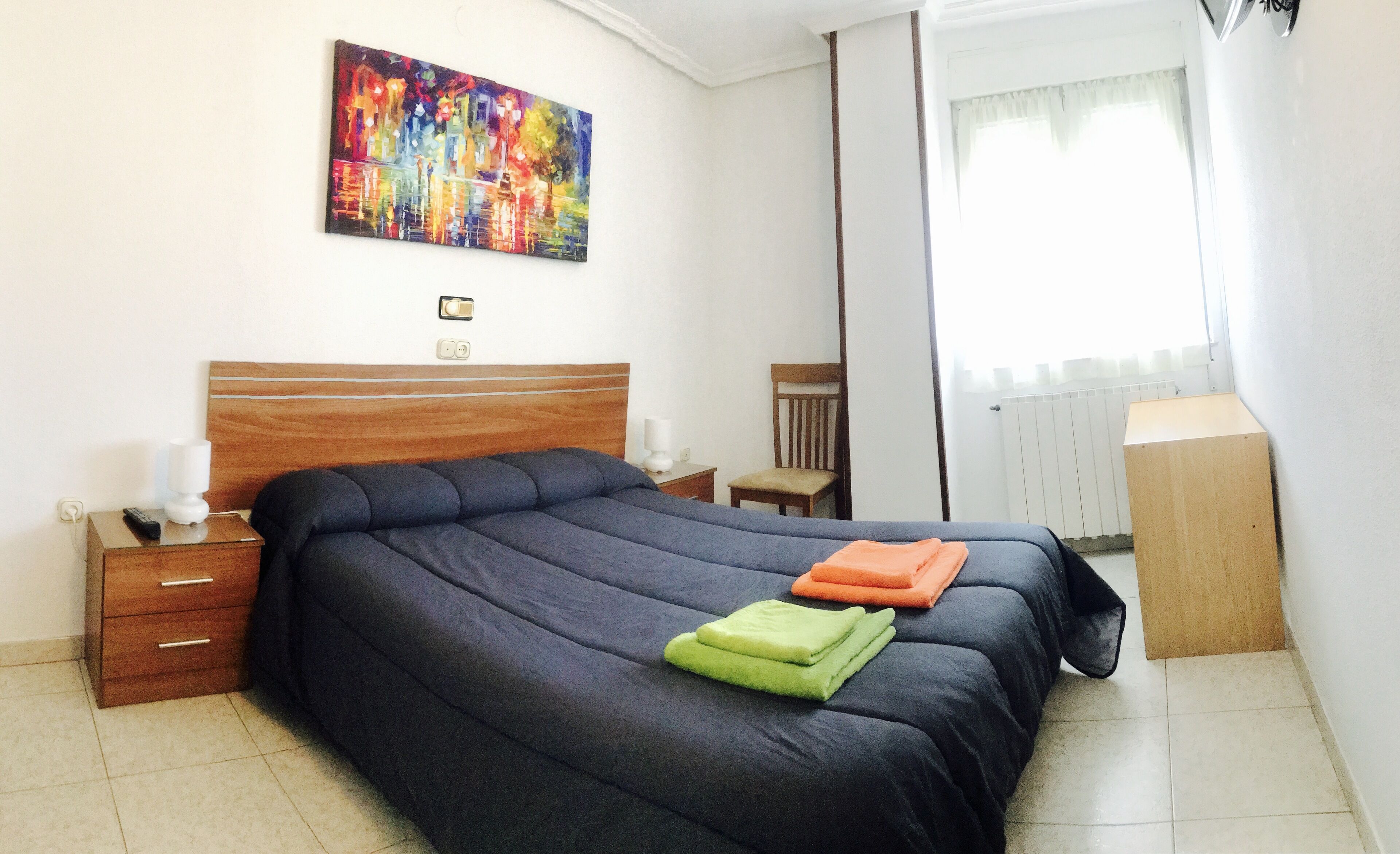 Een netjes opgemaakt slaapkamer met een groot bed, een nachtkastje met een lamp, een stoel en een radiator.