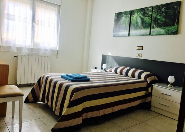 A hotel room with a bed, bedside table, lamp, and a painting of a forest on the wall. - Hostal Sol Naciente (León)