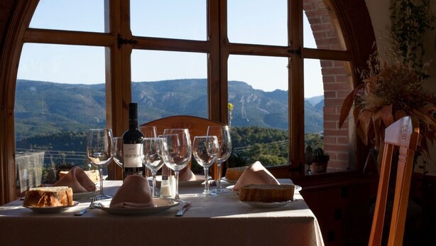 A table set for dining with a wine bottle, glasses, bread, and a view of mountains through large windows.
