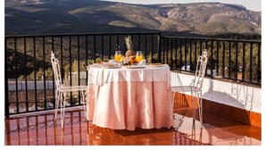 A table with a tablecloth and two chairs on a balcony with a mountain view.