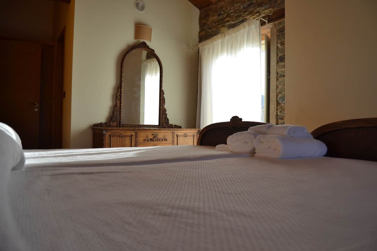 Een net opgemaakt bed met witte lakens, een houten dressoir met spiegel en een raam met dunne gordijnen.