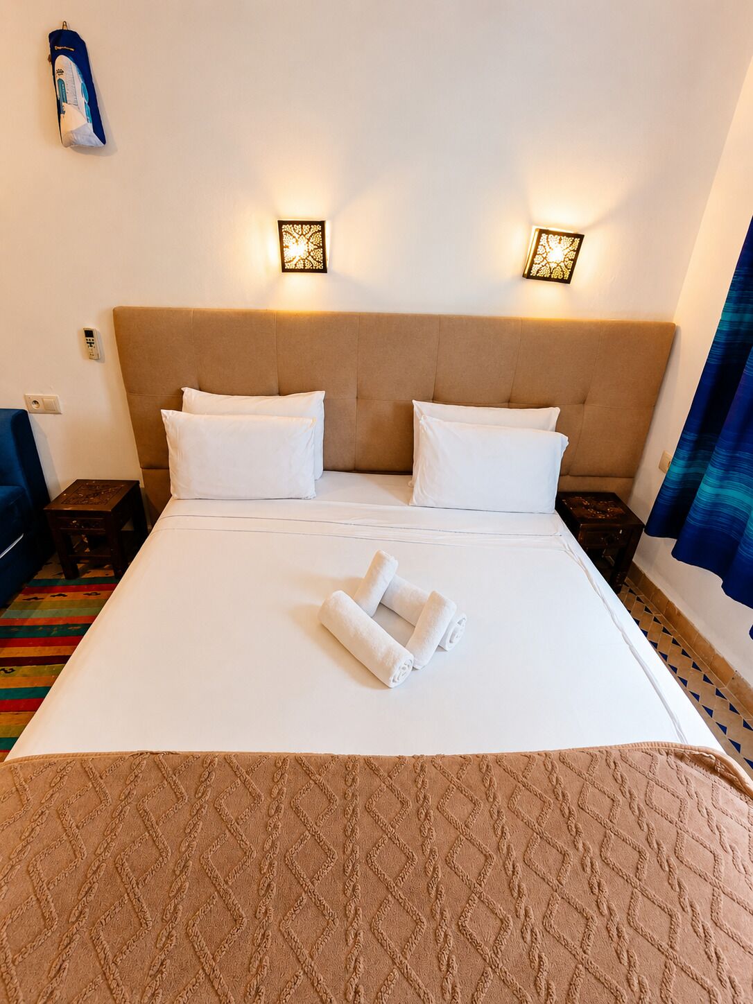 A hotel room with a large bed, two bedside lamps, a control panel, a blue chair, and a colorful rug.