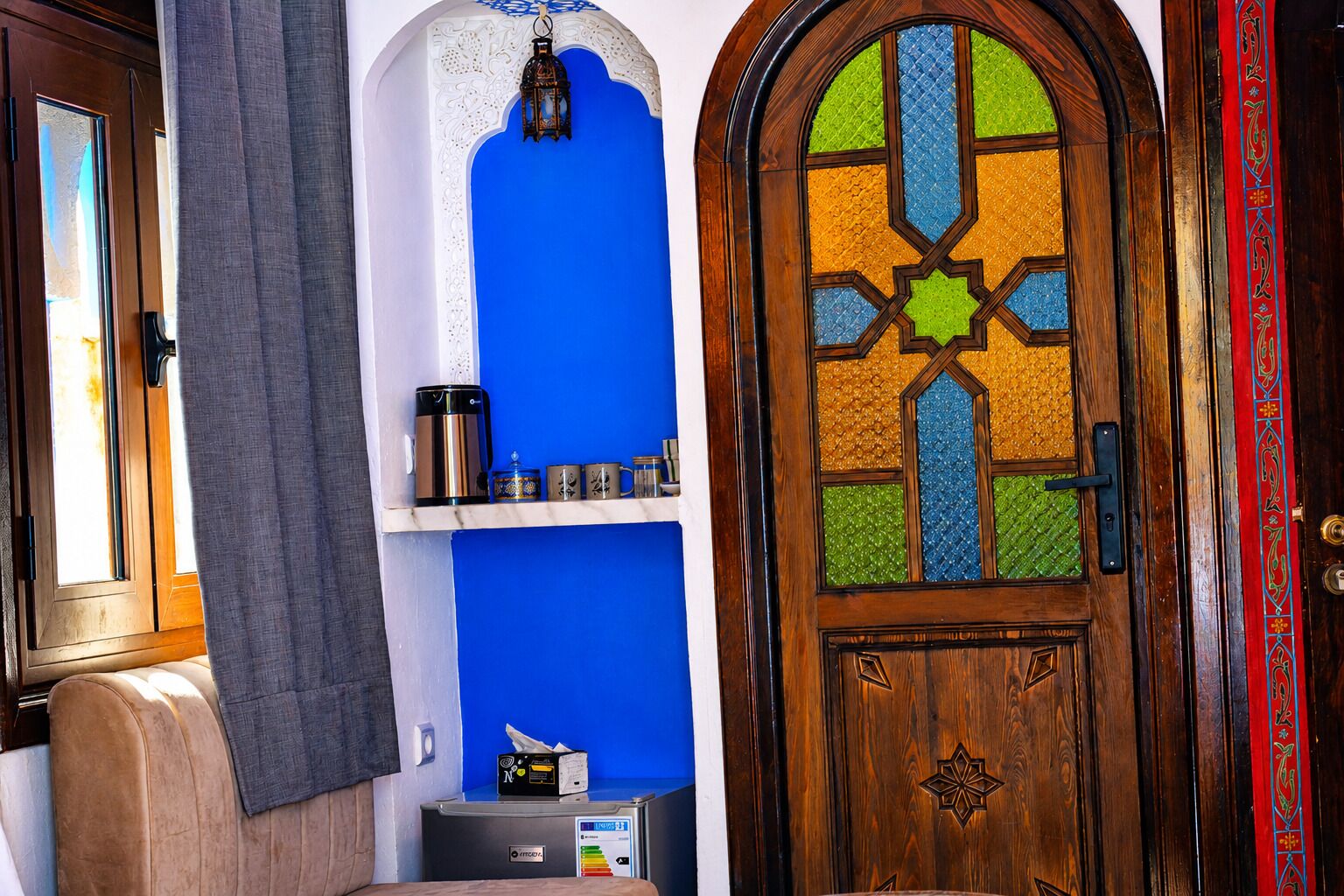 Une pièce avec un mur bleu, une porte en bois ornée d’un motif géométrique et une étagère sur laquelle se trouvent une bouilloire et des récipients.