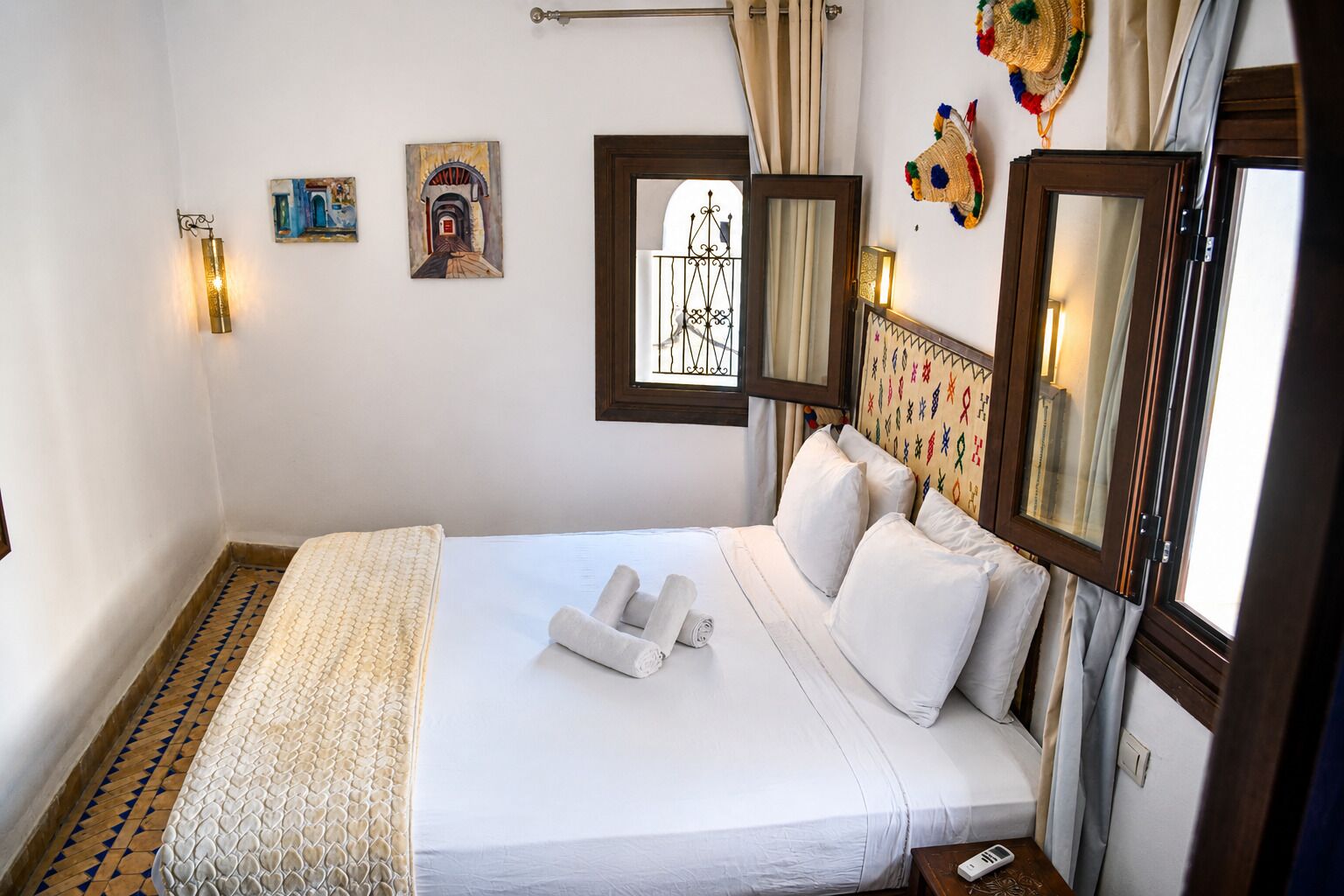 Un lit bien fait, avec du linge de lit blanc et des serviettes pliées, une table de chevet avec une lampe, un miroir fixé au mur et deux tableaux accrochés au mur.