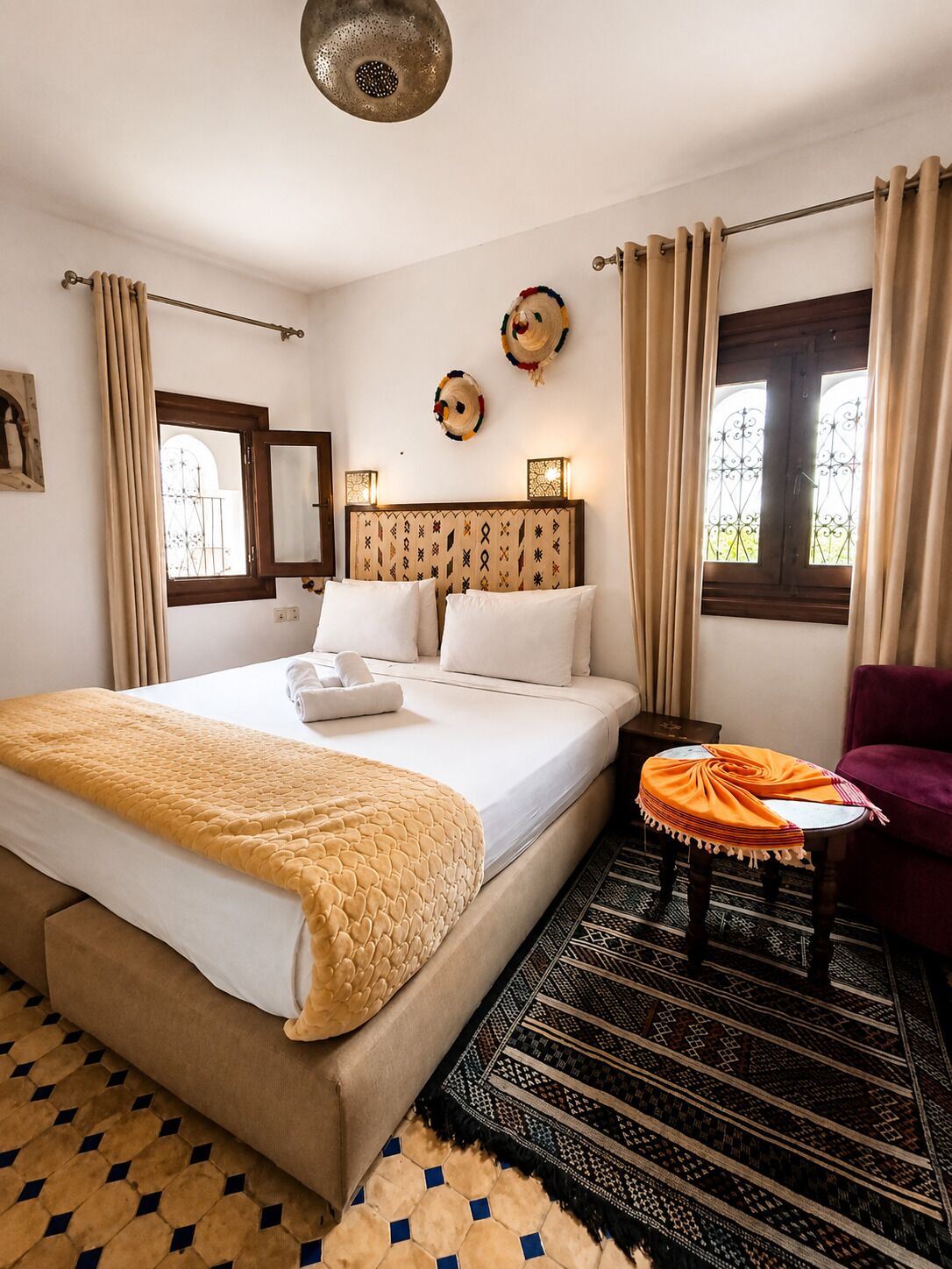 Una habitación de hotel con cama, una silla, una mesa, un tapete estampado y una ventana con cortinas.