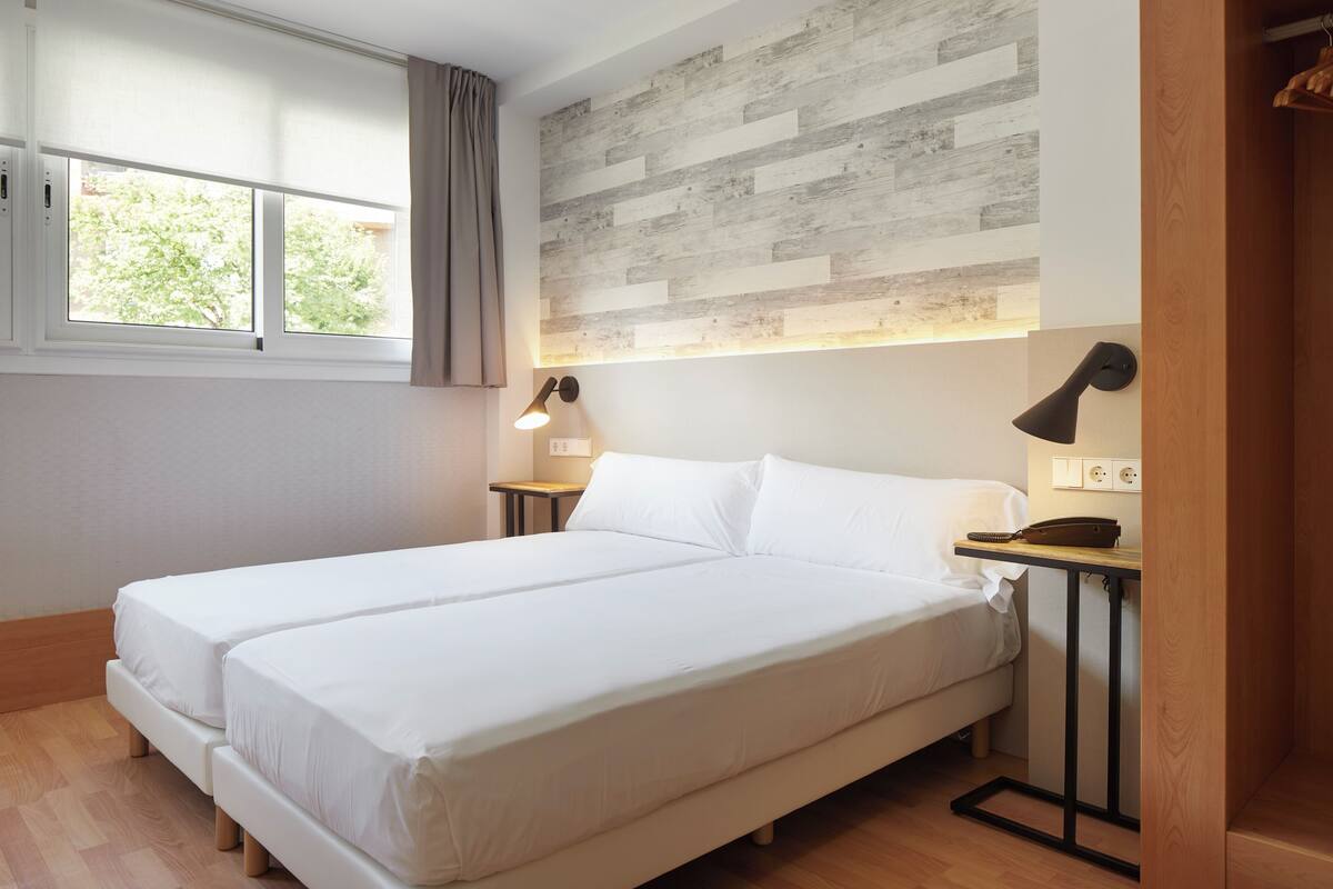 Un dormitorio con dos camas, un panel de madera en la pared, una ventana con cortinas y lámparas de noche.