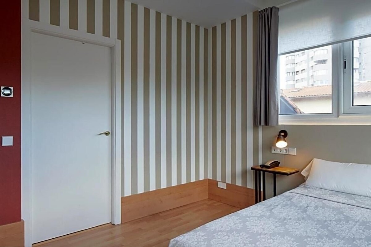 A room with a bed, a window with curtains, a bedside table with a lamp, and a door.