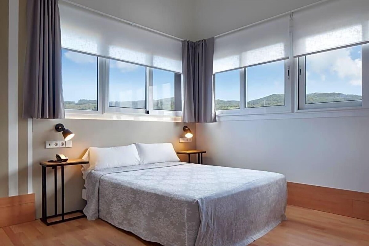 A bedroom with a bed, two bedside tables, two wall-mounted lamps, and large windows with curtains.