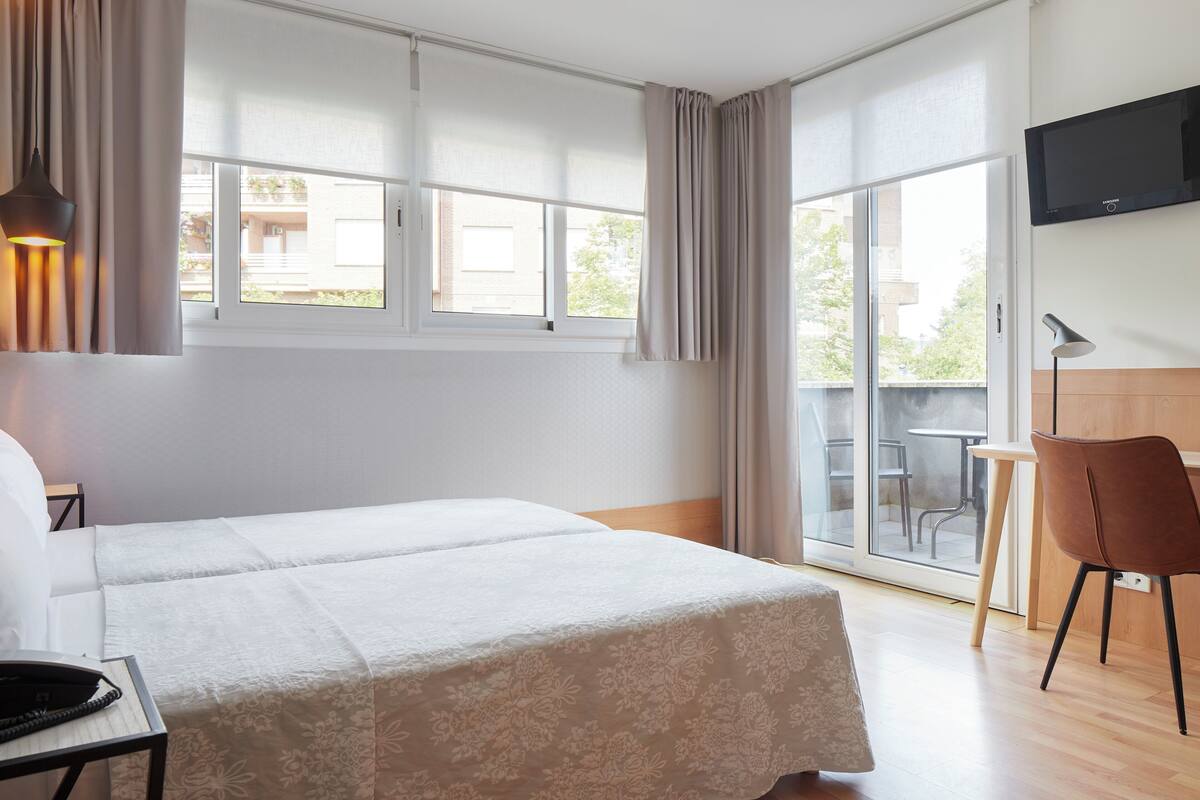 Una habitación de hotel con cama, escritorio con teléfono, silla, televisor en la pared y balcón con mesa y sillas.