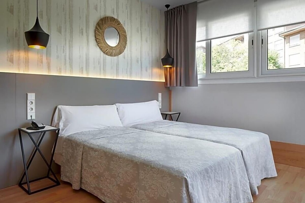 A double bed with a light-colored bedspread, two bedside tables with lamps, a wall-mounted mirror, and a window with curtains.