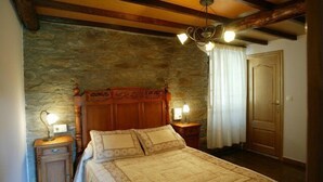 Een slaapkamer met een stenen muur, houten balken in het plafond, een bed met hoofdeinde, bedlampen en een deur.