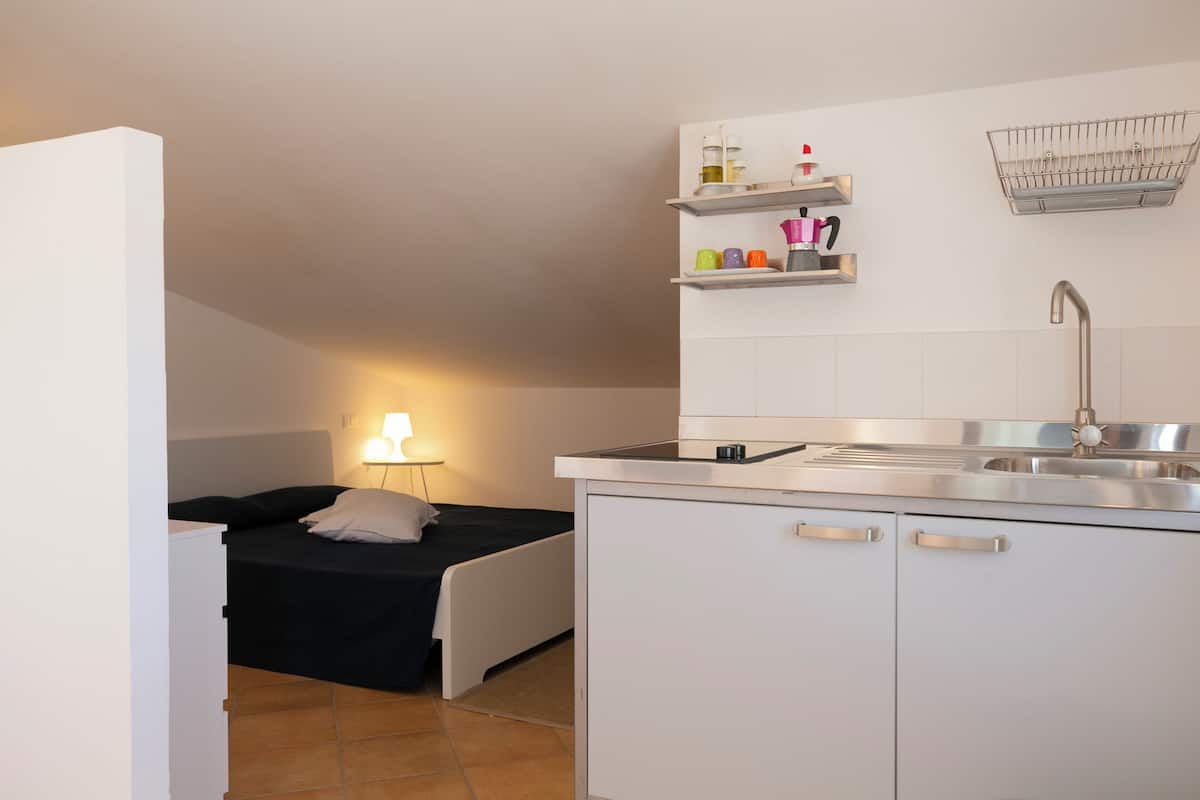 A compact kitchen with a sink, cabinets, and a small countertop. There is a bed with a nightstand and a lamp in the background.