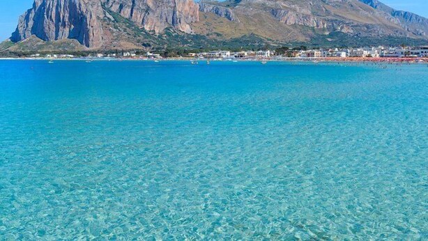 A coastal town with clear turquoise waters, rocky mountains in the background, and a beach with buildings.