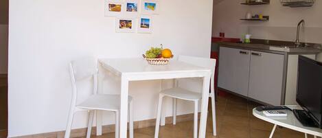 Une petite cuisine moderne avec des meubles blancs, une table à manger avec deux chaises et un panier de fruits posé sur la table.