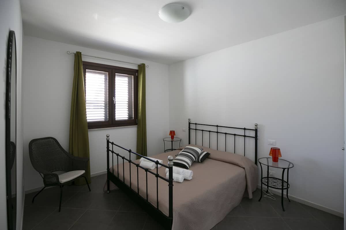 A bedroom with a bed, a chair, a small table, a window with curtains, and a ceiling light.