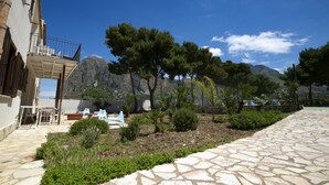 A courtyard with stone flooring, a small garden with various plants, and a mountain in the background.