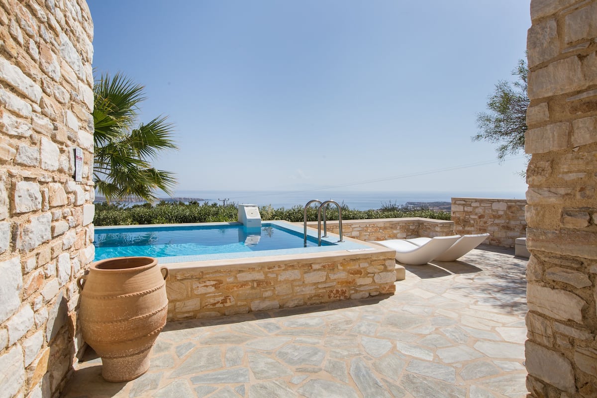 Un patio de piedra con piscina, sillas de descanso blancas y vistas al mar.