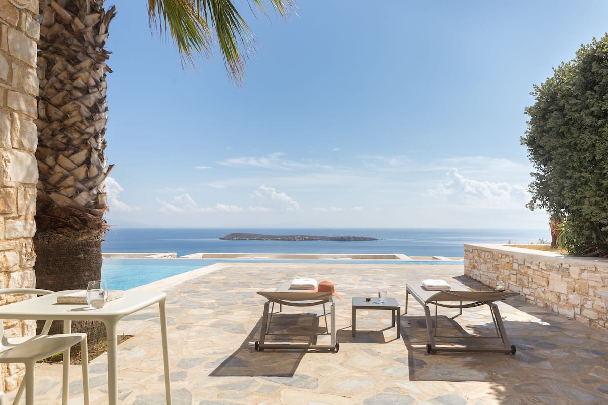 Un patio de piedra con dos sillones de descanso y una mesa, con vista a una piscina y al mar.