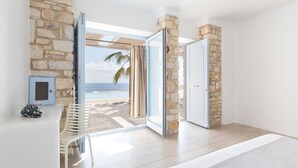 A room with a stone wall, a sliding glass door leading to a balcony with a view of the beach and palm trees. - Calme Boutique Hotel Paros- Adults Only (Paros)