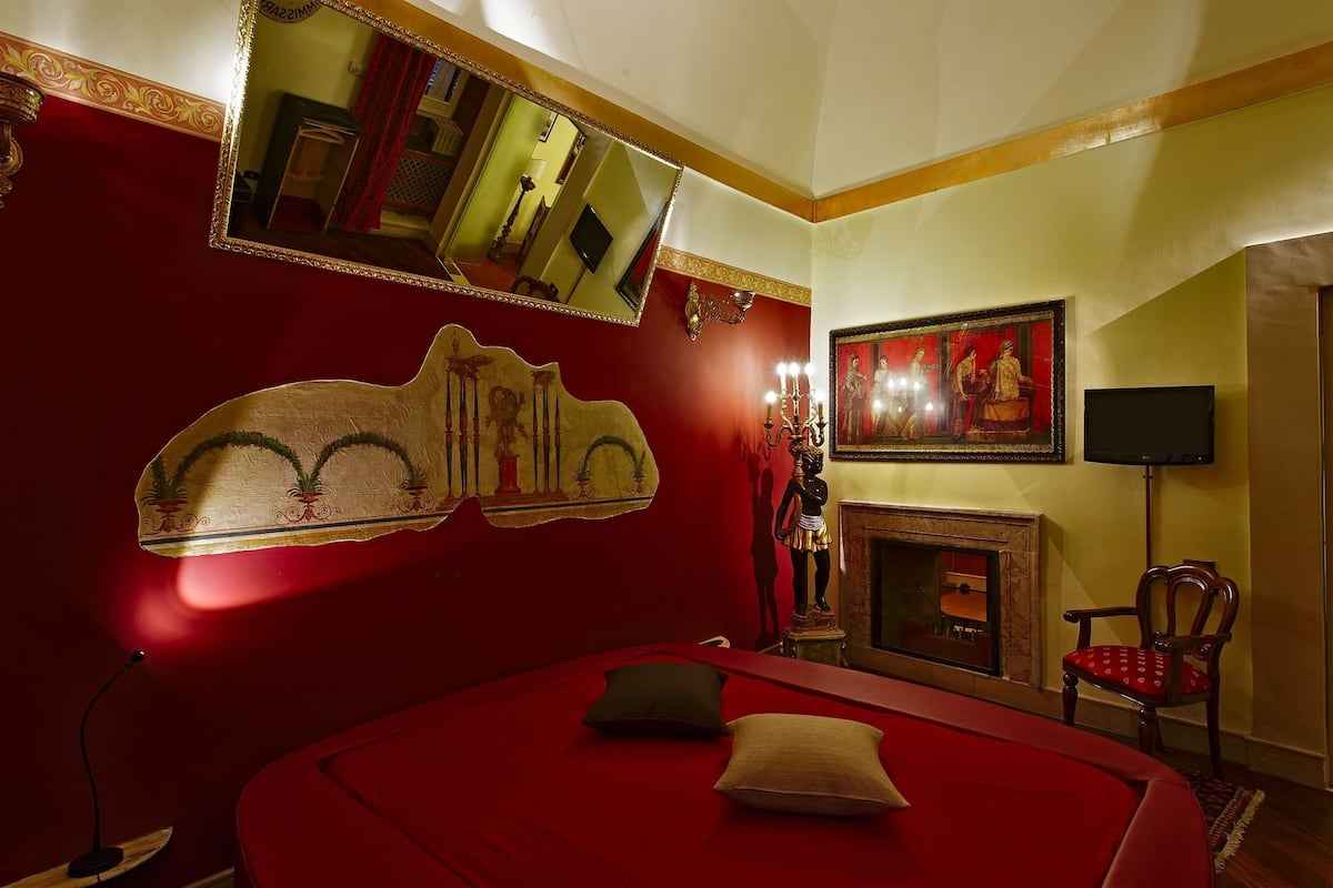 Una habitación con una cama roja, un espejo grande, una chimenea y una silla.