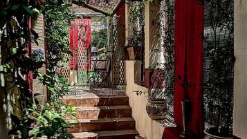 Un estrecho pasillo exterior con escalones que llevan a una zona cubierta, rodeado de plantas en macetas y un paño rojo.