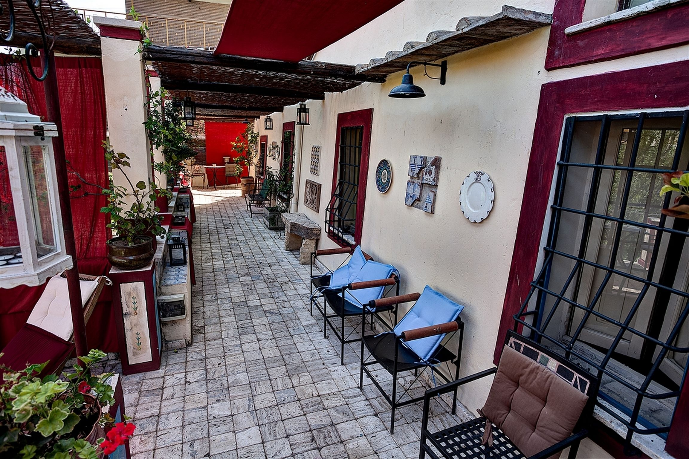 A narrow courtyard with a tiled floor, a red awning, and several chairs. There are potted plants and a small table with a lamp.