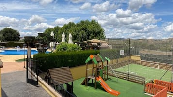 Un espacio exterior cercado con un parque infantil, cancha de tenis y piscina.