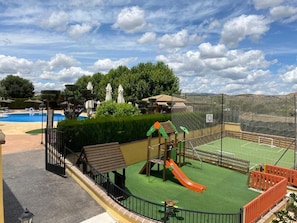 Un espacio exterior cercado con un parque infantil, cancha de tenis y piscina.