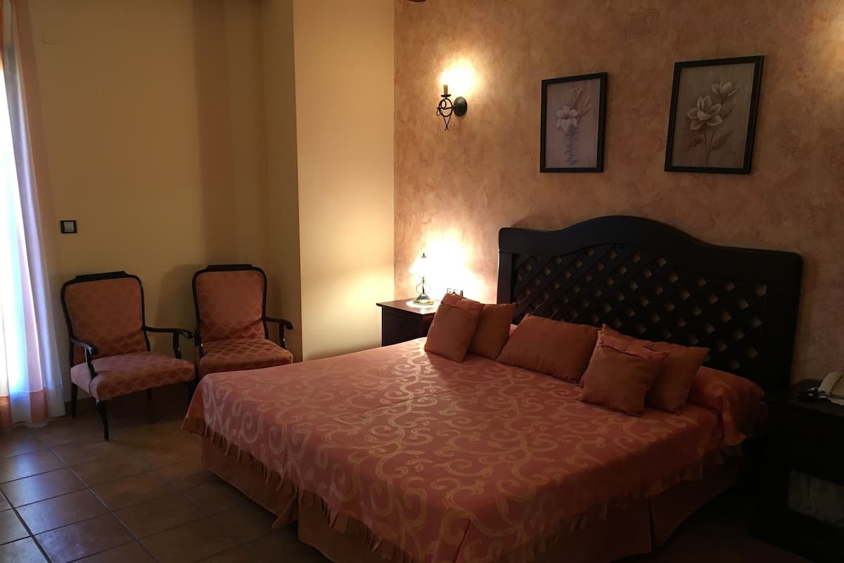A bedroom with a large bed, two chairs, a nightstand, and a wall-mounted lamp.
