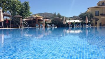 A clear blue pool with lounge chairs and umbrellas, surrounded by buildings and trees.