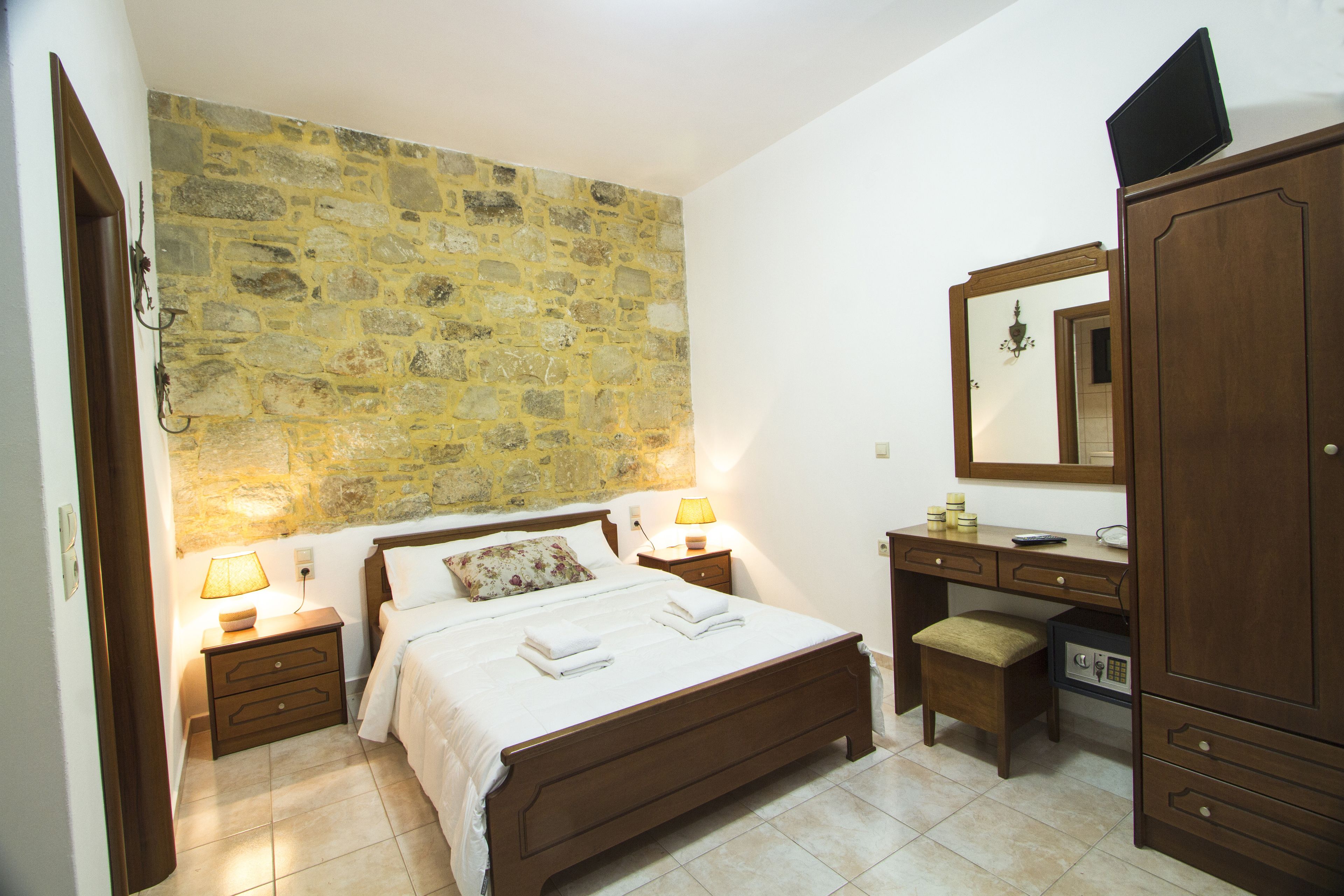 A bedroom with a stone wall, a wooden bed, bedside tables with lamps, a desk with a mirror, and a safe.