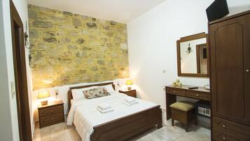 A bedroom with a stone wall, a wooden bed, bedside tables with lamps, a desk with a mirror, and a safe.