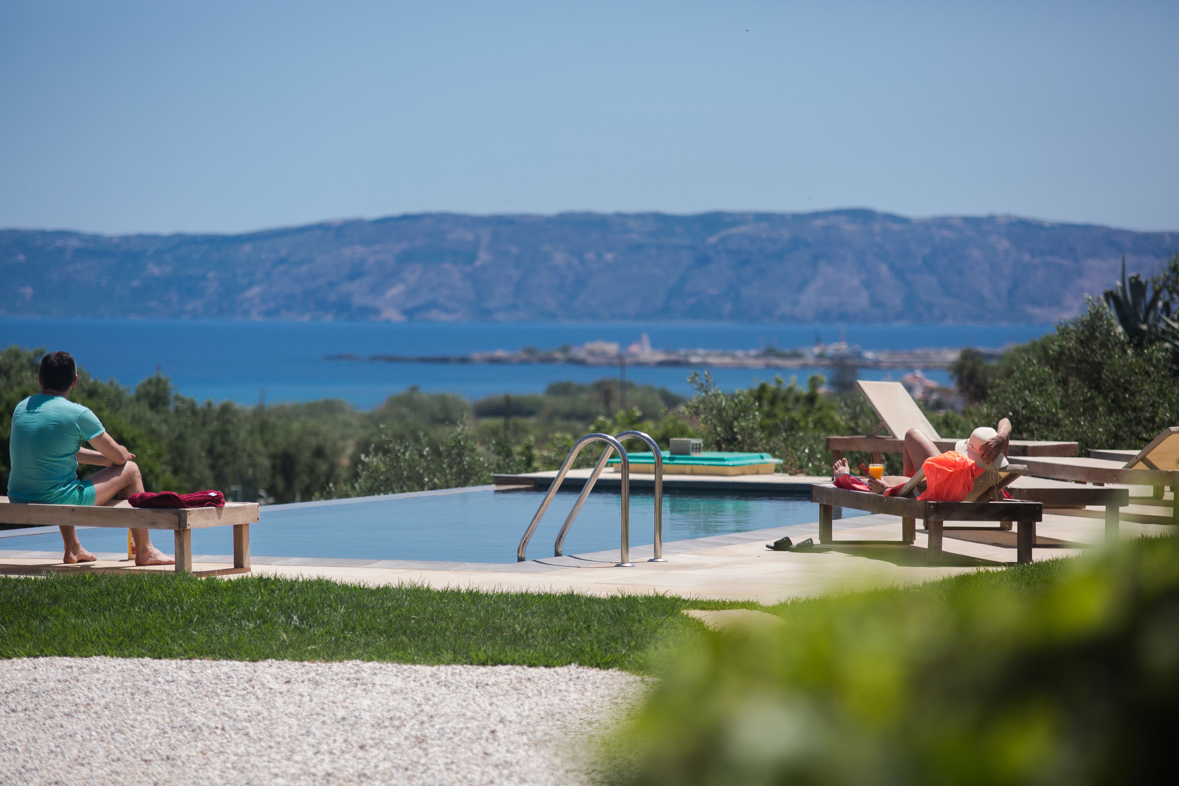 A poolside area with a view of the sea and hills, including lounge chairs and a pool.