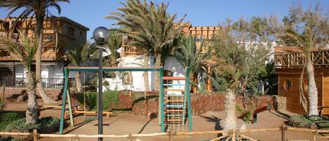 A playground with swings and climbing structures, surrounded by palm trees and a building with a traditional architectural style.