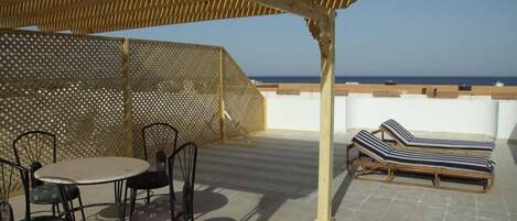 A covered outdoor seating area with a table and chairs, a lounge chair, and a view of the sea.