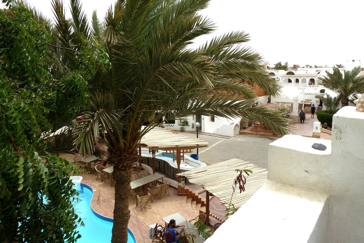 A pool area with palm trees, outdoor seating, and a view of a white building with arches.