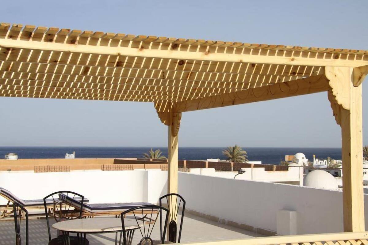 A covered outdoor seating area with a view of the sea, featuring a pergola structure and a table with chairs.