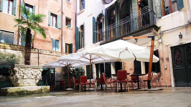 An outdoor seating area with tables and chairs under umbrellas, a palm tree, and a stone planter. - Residenza Della Spada (Venice)