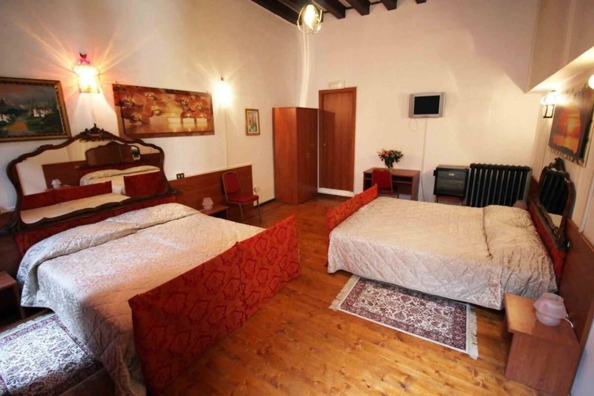 A hotel room with two beds, wooden flooring, a radiator, a TV, and artwork on the walls.