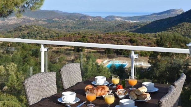 A table set with breakfast items, including croissants, bread, and juice, overlooking a scenic landscape with hills and a pool. - Hôtel Résidence U Nebbiu (Olmeta-di-Tuda)