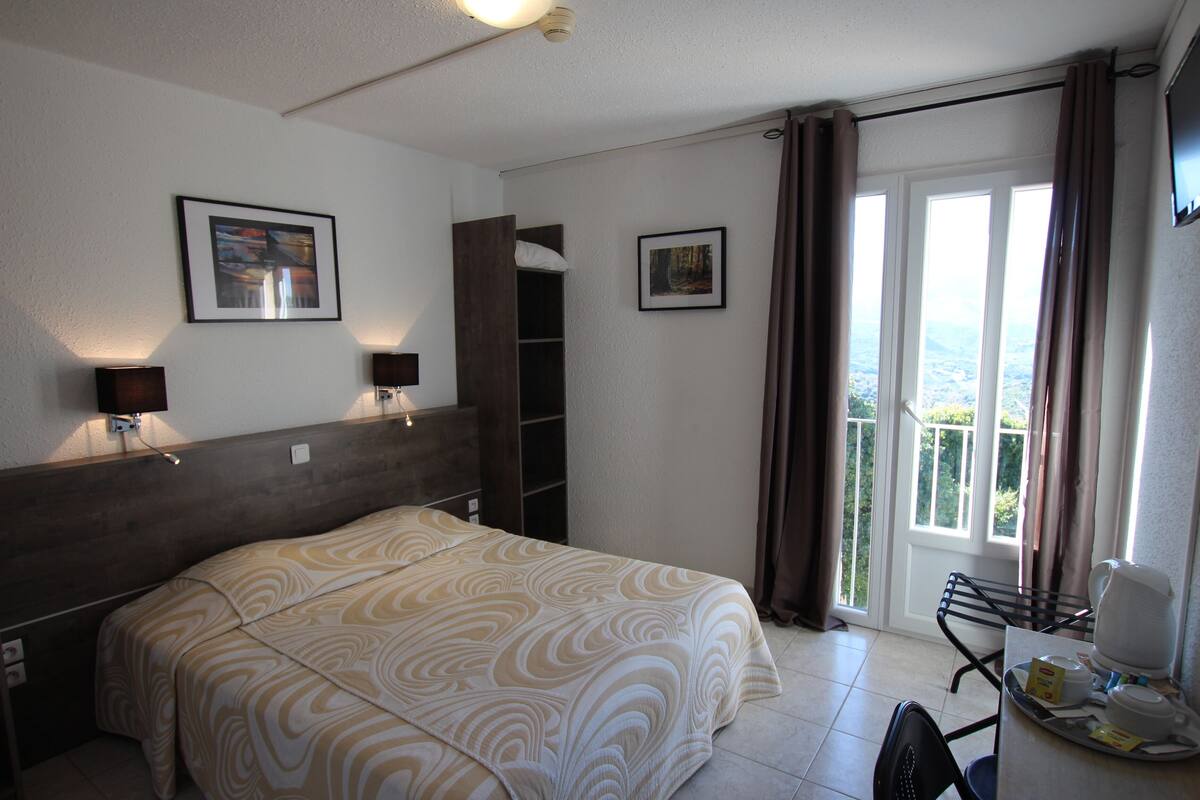 Une chambre d’hôtel comprenant un lit, un bureau avec une chaise, une fenêtre avec des rideaux et deux lampes fixées au mur.