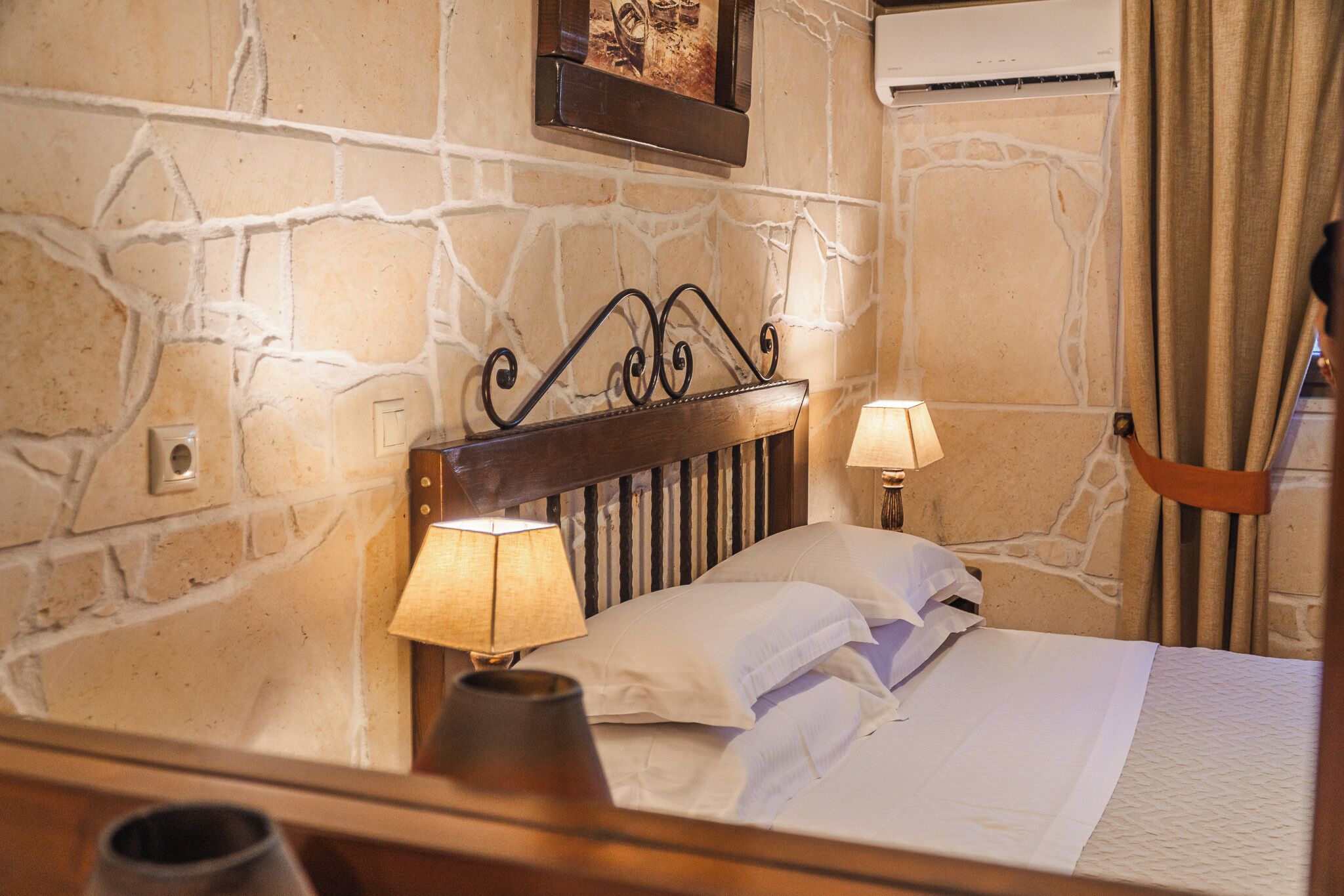 Habitación de hotel con pared de piedra, cabecera de madera, cama con ropa blanca, dos lámparas de noche y un equipo de aire acondicionado.